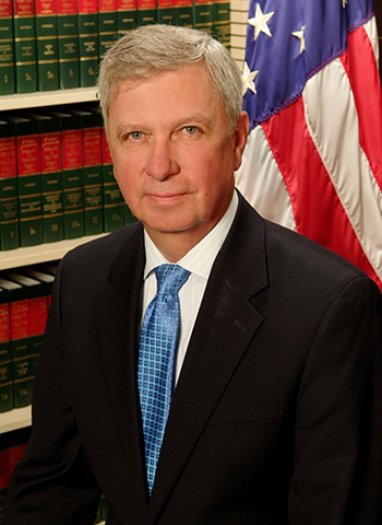 Professional headshot of a man in a suit with an American flag background.